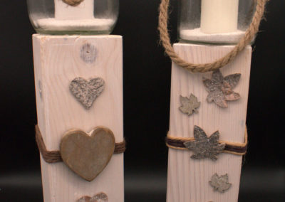 Etwas moderner schlicht in weiß gehalten Windlichtglas mit weißer Kerze. Holzbalken mit Herzen und Blättern dekoriert.